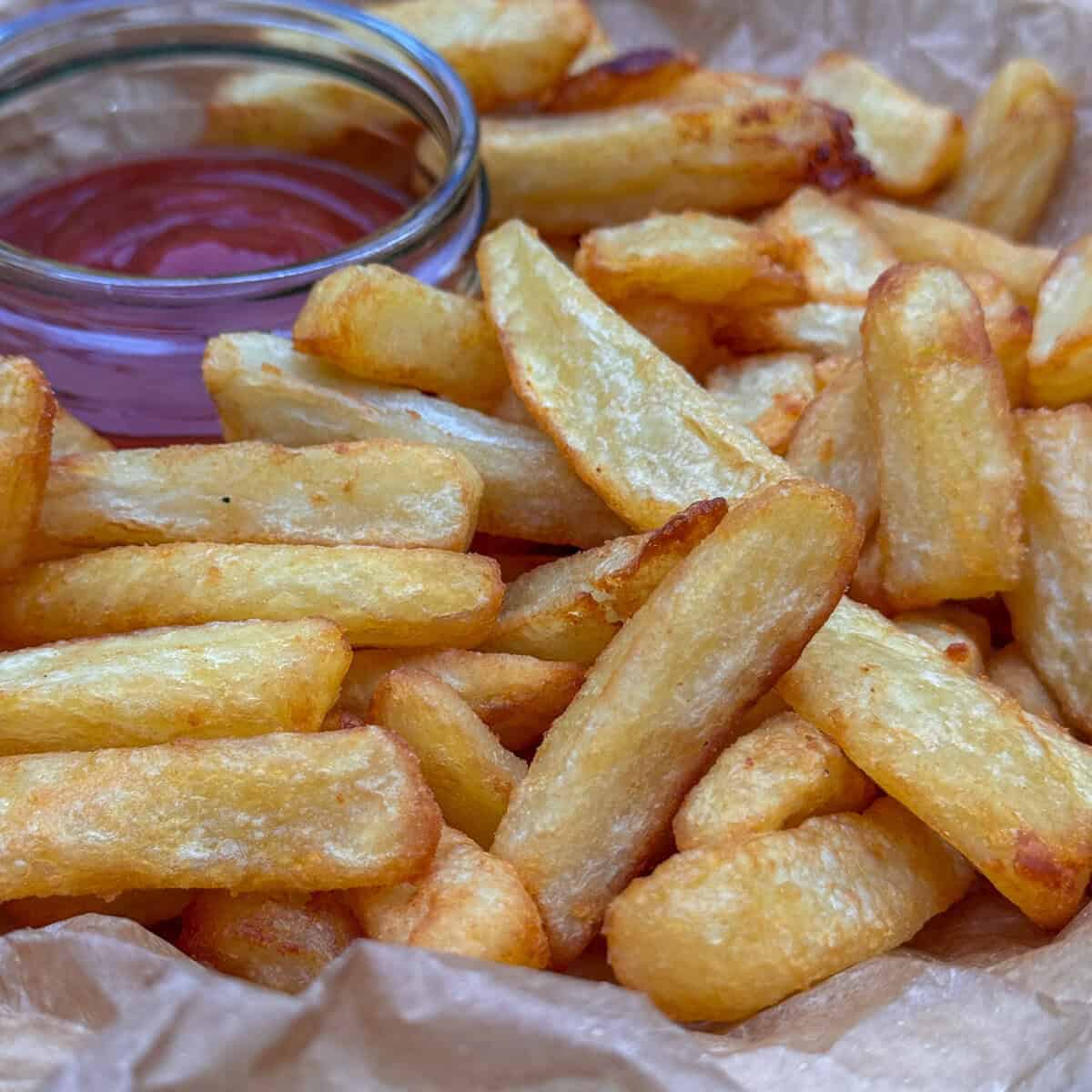 A close-up of golden crispy homemade chips on parchment paper with a glass container of red ketchup in the background.