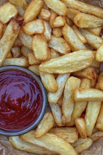 A pile of golden, crispy homemade chips on parchment paper, with a glass bowl of ketchup placed to the side for dipping.