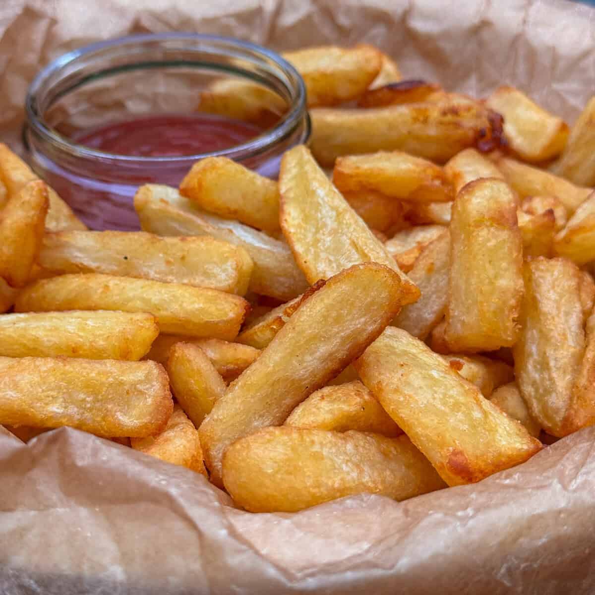 A pile of golden, crispy homemade chips served on parchment paper with a small glass bowl of ketchup in the background.