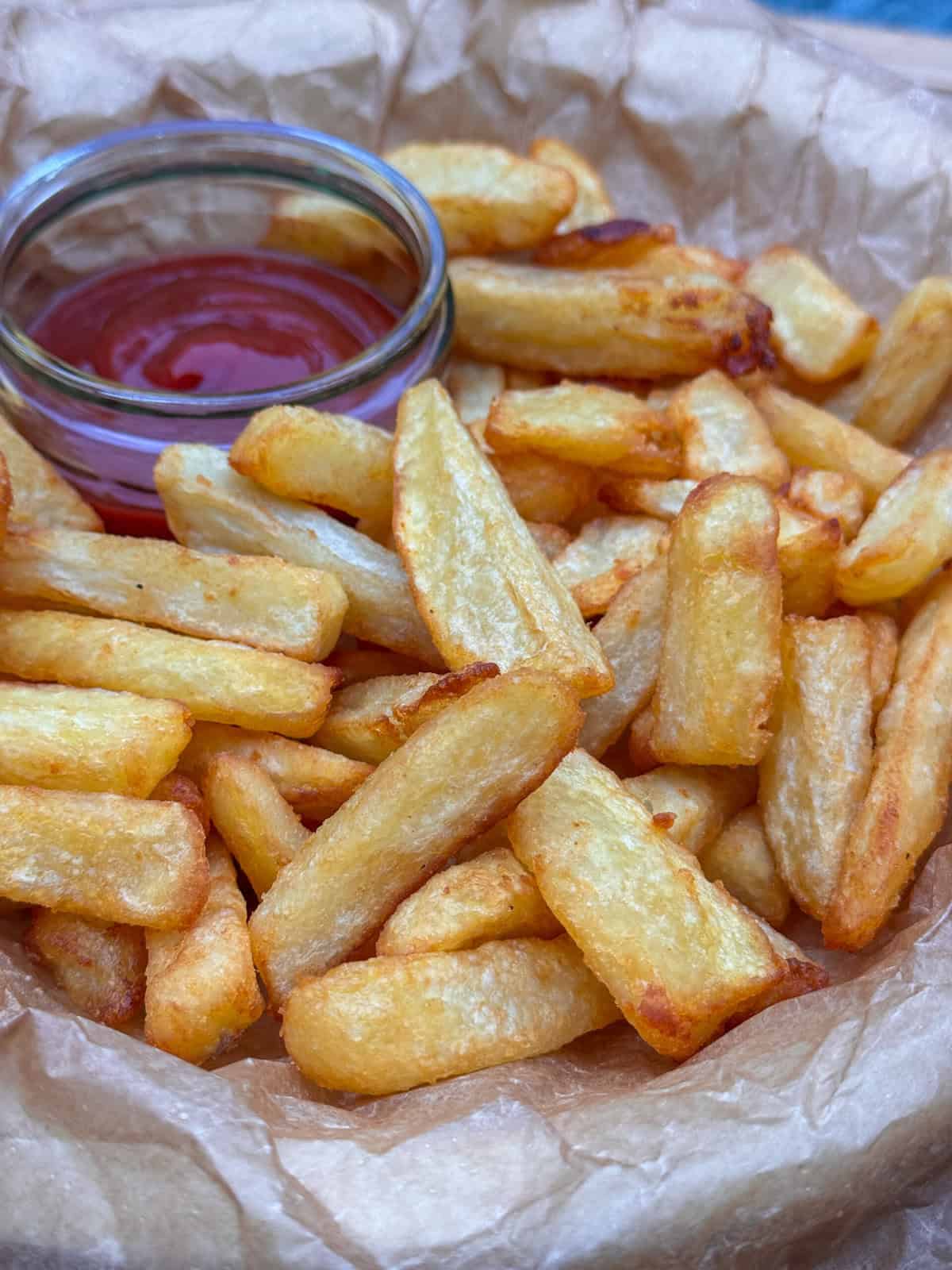 A basket lined with parchment paper filled with golden, crispy homemade chips. A small glass container of ketchup sits in the corner for dipping.