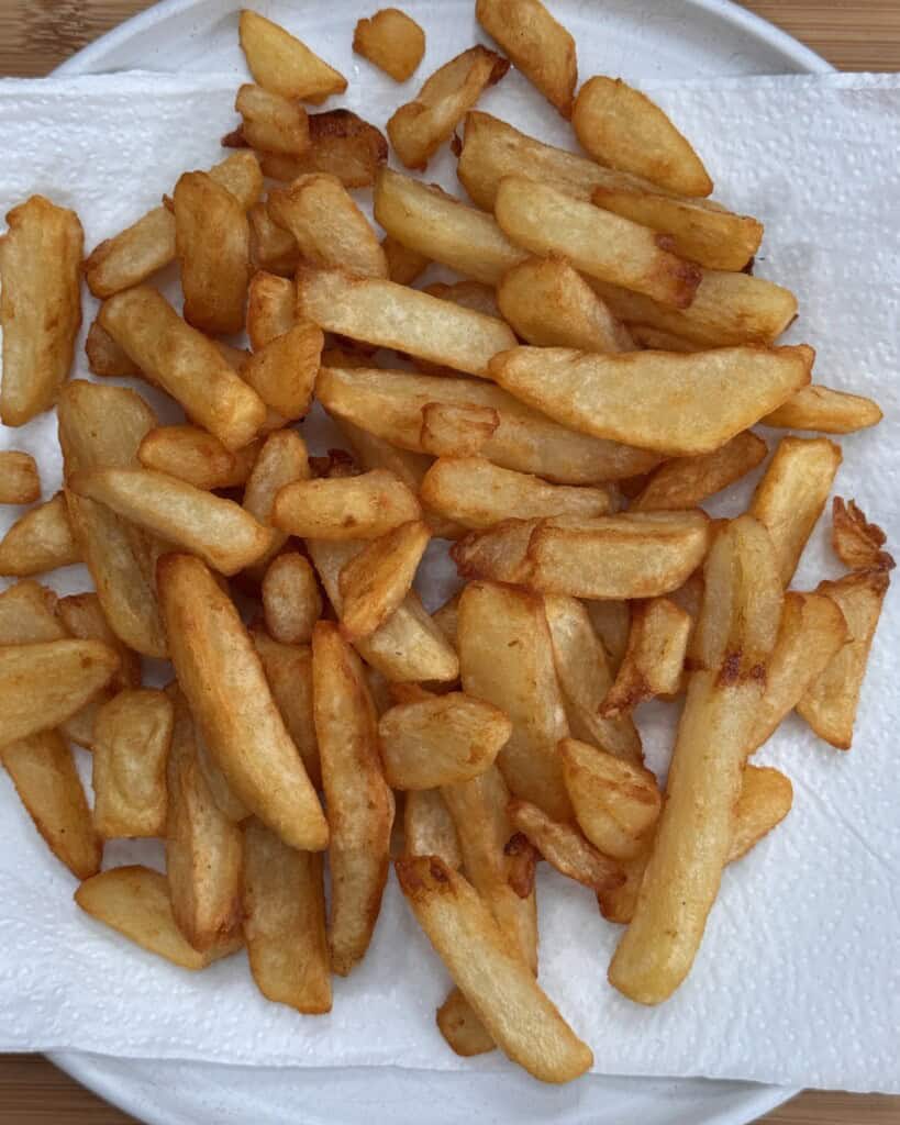 Golden, crispy homemade chips are spread out on a white paper towel, which is placed on a white plate. The fries appear freshly cooked, with a slight sheen and some variation in size and shape.