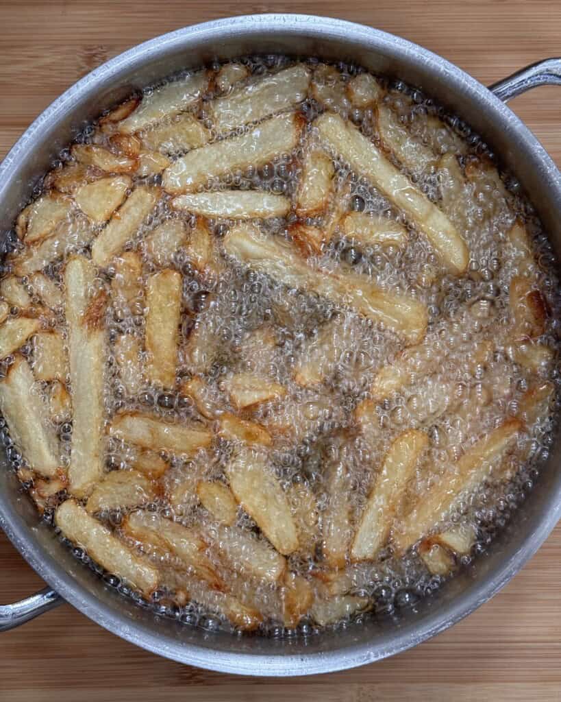 A pot filled with hot oil frying golden brown crispy homemade chips, with bubbles forming around the fries as they cook. The pot is placed on a wooden surface.