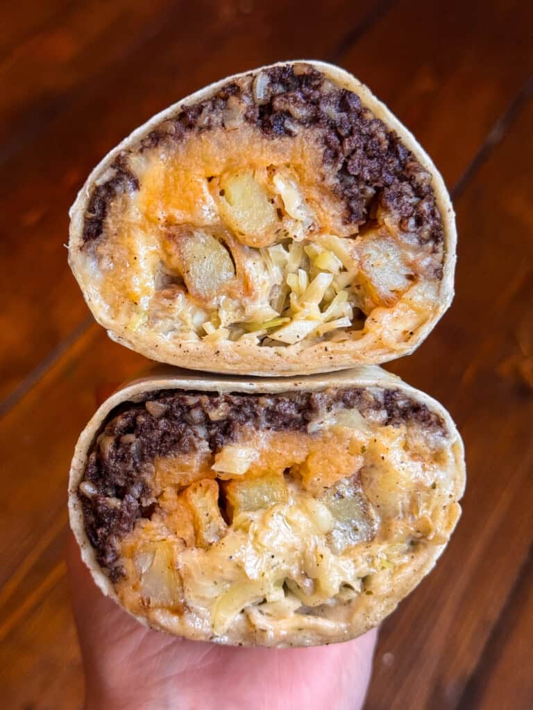 A hand holds a burrito cut in half, revealing layers of seasoned ground beef, melted cheese, potatoes, and shredded lettuce inside a tortilla. The background is a wooden surface.