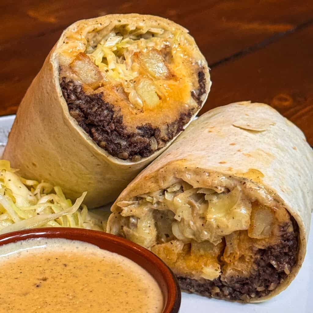 A haggis burrito cut in half, showing fillings of black beans, potatoes, cheese, and shredded lettuce. A side of shredded cabbage and a small bowl of creamy sauce are on the plate. The background is a wooden table.