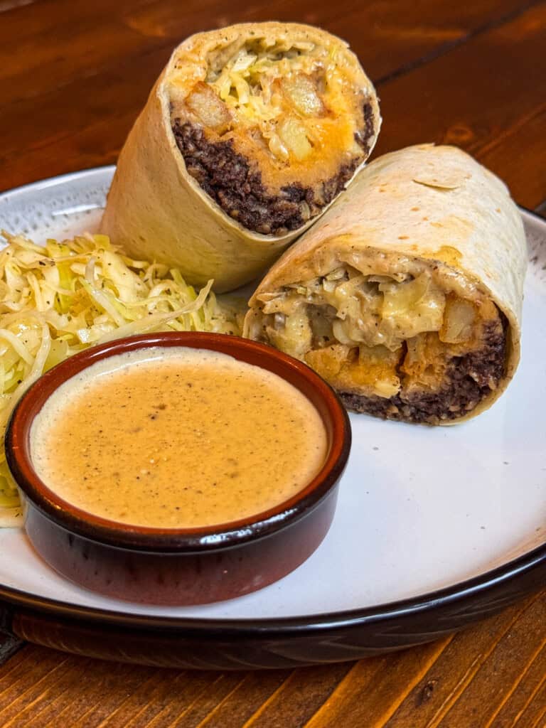 A burrito cut in half shows black beans, potatoes, and cheese inside. The plate also has a serving of shredded cabbage and a small bowl of creamy dipping sauce. The food is on a white plate on a wooden table.