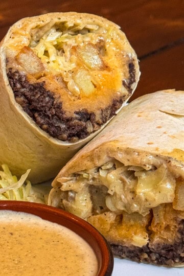 A haggis hoagie (haggis burrito) cut in half, showing fillings of black beans, potatoes, cheese, and shredded lettuce. A side of shredded cabbage and a small bowl of creamy sauce are on the plate. The background is a wooden table.