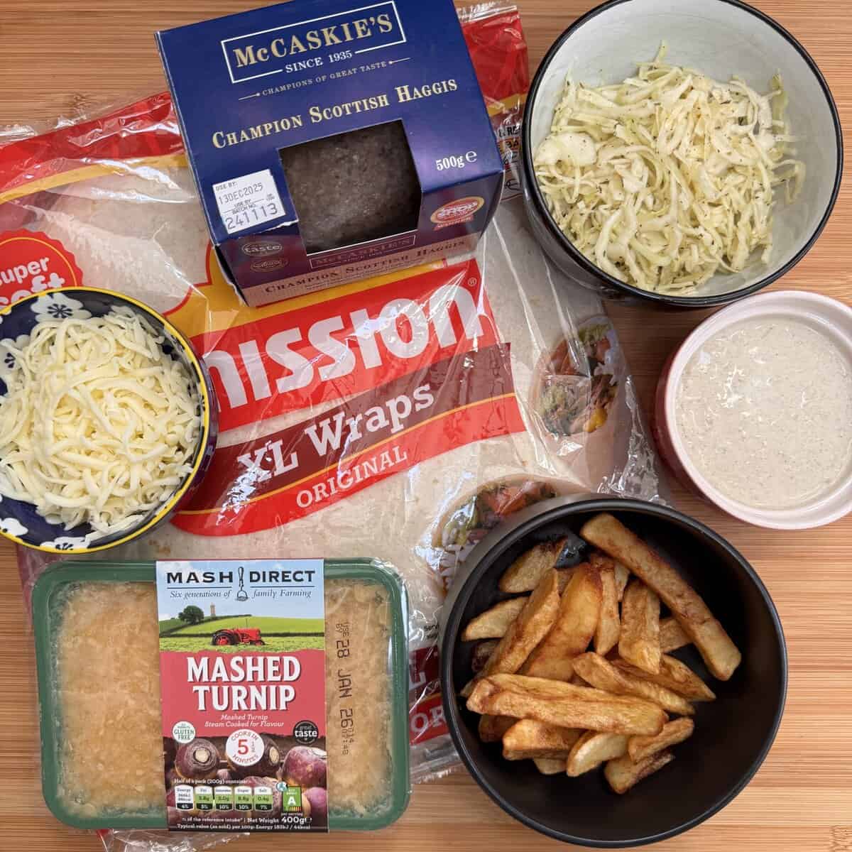 A selection of food items on a wooden surface: XL tortilla wraps, a pack of Scottish haggis, mashed turnip, grated cheese, fries, shredded cabbage in a bowl, and a bowl of creamy sauce.