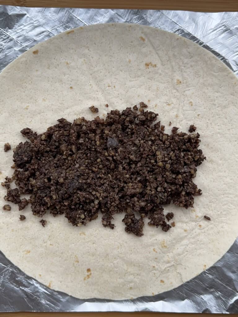 A flour tortilla on a sheet of foil with a filling of finely ground, dark-colored cooked mixture spread across the center, ready to be wrapped.