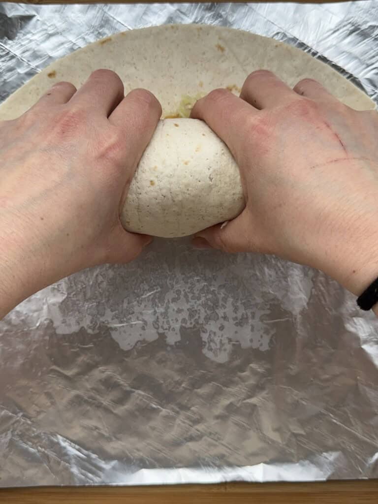Two hands are seen folding a tortilla wrap filled with food on a sheet of aluminum foil. The wrap is being rolled tightly, with the edges pinched in.