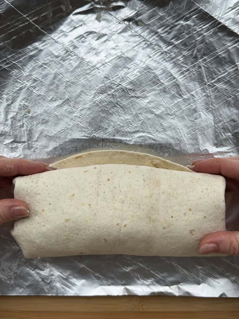 Two hands are wrapping a tortilla burrito on a sheet of aluminum foil, preparing it for folding or wrapping.
