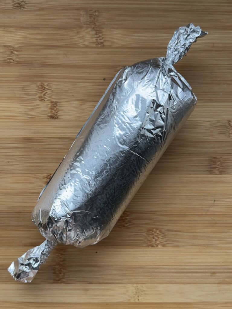 A log-shaped object wrapped tightly in aluminum foil, twisted closed at both ends, resting on a wooden surface.
