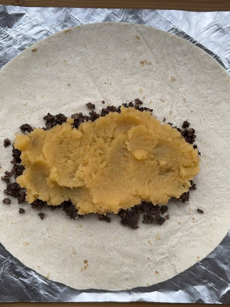 A flour tortilla on foil topped with a layer of cooked ground beef and a layer of mashed potatoes, ready to be wrapped.
