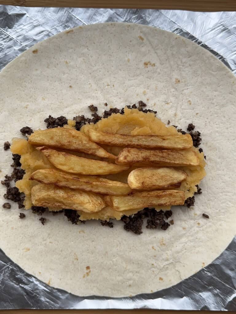 An open tortilla on foil topped with a layer of crumbled meat, mashed yellow plantains, and several fried potato wedges, ready to be wrapped.