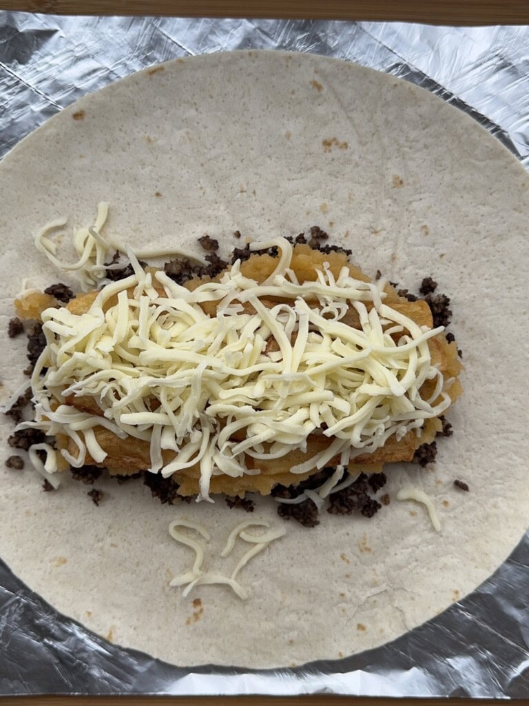 A flour tortilla on foil topped with ground beef, hash brown patties, and shredded cheese, ready to be wrapped.