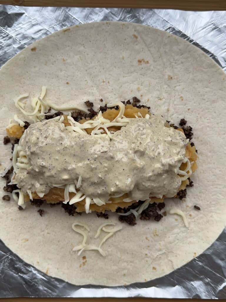 A flour tortilla topped with shredded cheese, ground meat, hash brown patties, more cheese, and a generous amount of creamy sauce, all placed on foil and ready to be wrapped.