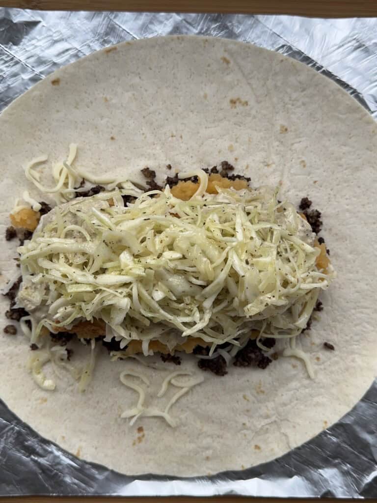 A flour tortilla on foil is filled with ground beef, shredded cheese, and a generous amount of seasoned coleslaw, ready to be wrapped.
