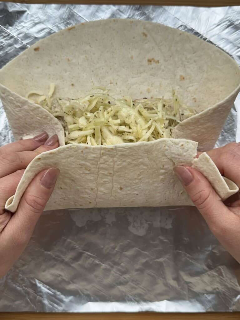 Close-up of hands folding a tortilla wrap filled with shredded cheese and other ingredients, positioned on a sheet of aluminum foil.