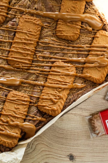 A tray of brownies topped with Biscoff biscuits and drizzled with Biscoff spread, with wrapped Biscoff biscuits on the side, on a wooden surface.