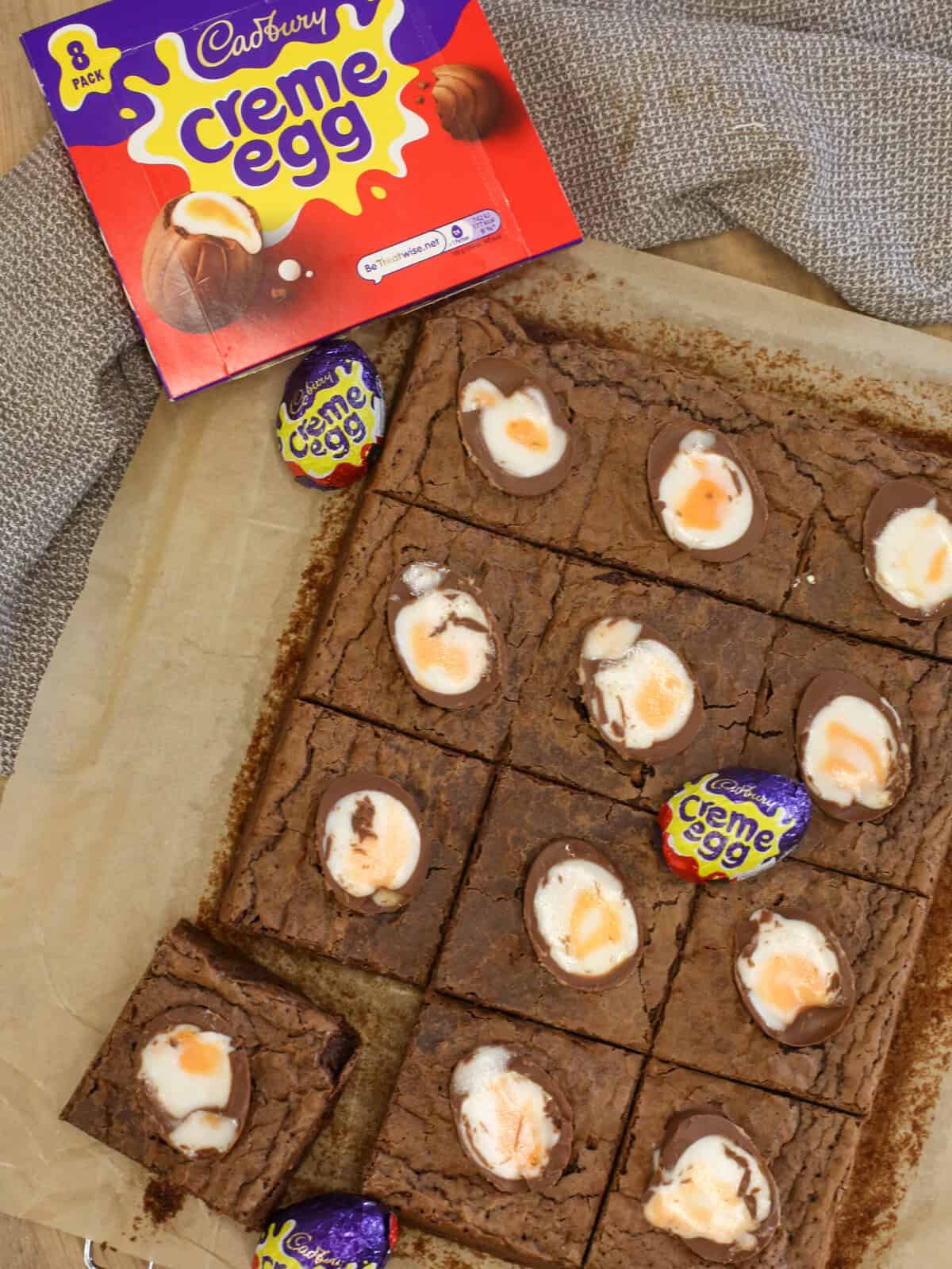 Brownies topped with halved Cadbury Creme Eggs on baking parchment, next to an opened box of Cadbury Creme Eggs and two wrapped eggs, all set on a textured brown cloth.