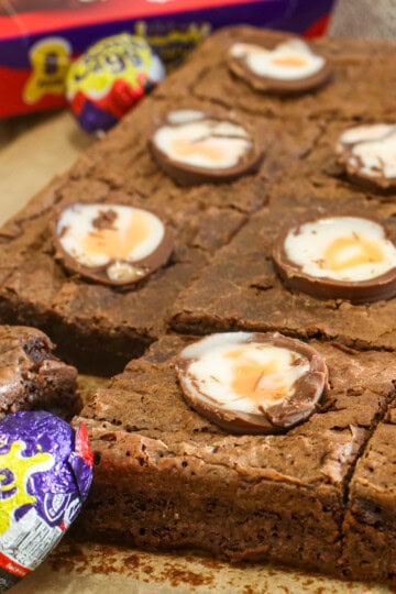 Close-up of chocolate creme egg brownies topped with halved Cadbury Creme Eggs, some whole Creme Eggs nearby, and a box of Creme Eggs in the background. The brownies are cut into squares and look rich and fudgy.
