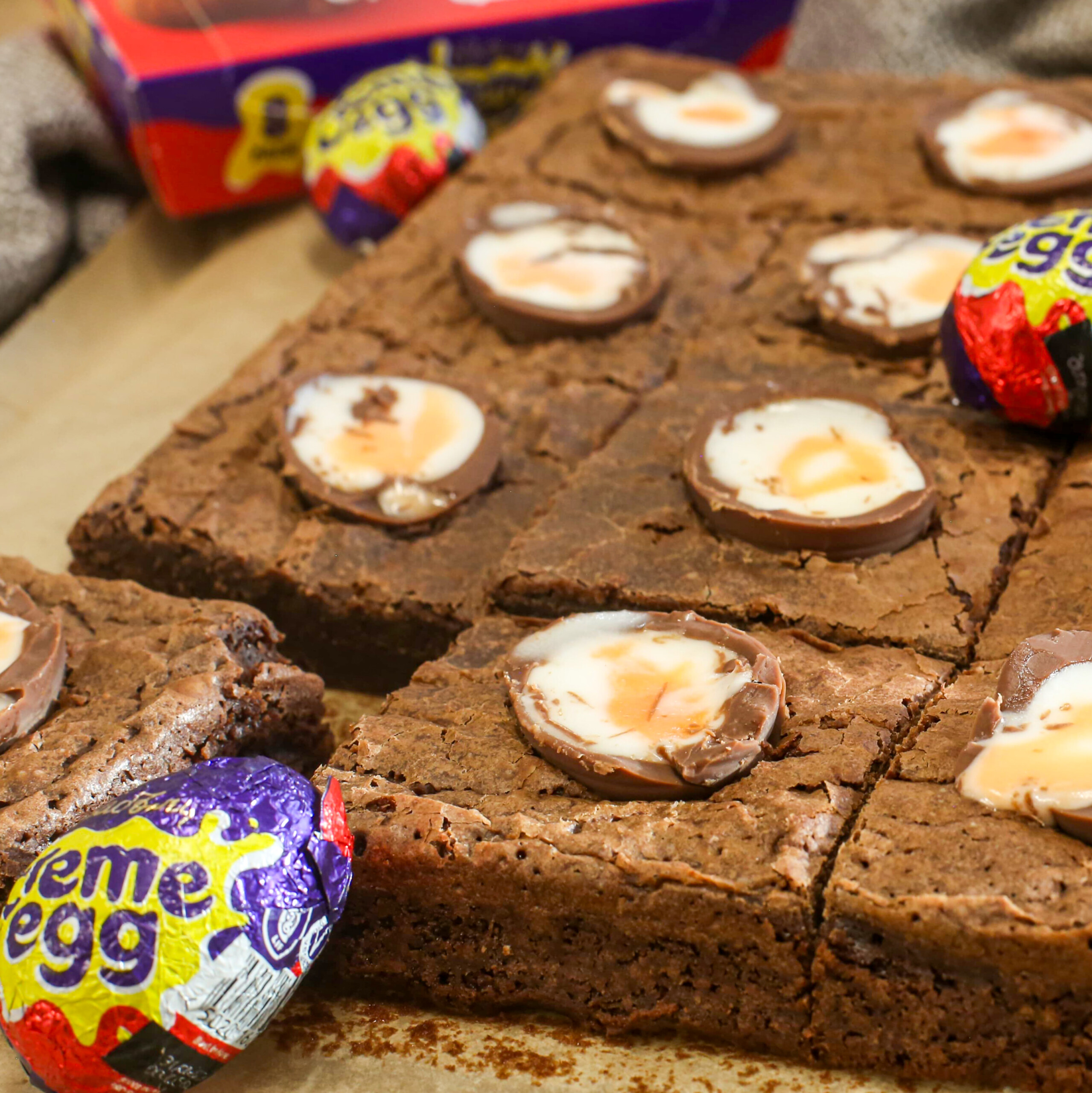 Close-up of chocolate creme egg brownies topped with halved Cadbury Creme Eggs, some whole Creme Eggs nearby, and a box of Creme Eggs in the background. The brownies are cut into squares and look rich and fudgy.
