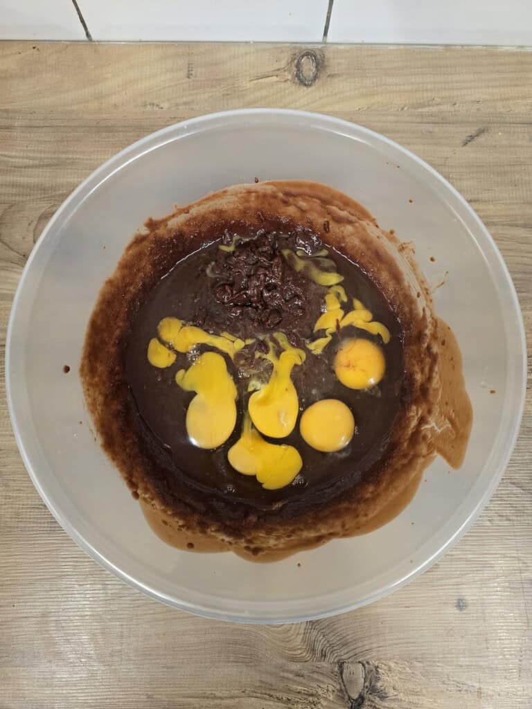 A clear mixing bowl on a wooden surface contains chocolate mixture and several cracked raw eggs, ready to be mixed together.