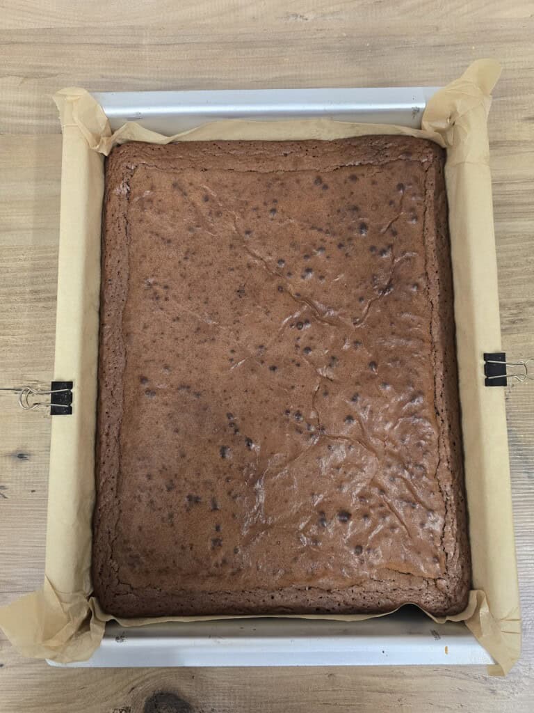 A large, freshly baked tray ofcreme egg brownies sits in a rectangular tin lined with baking paper, with metal clips holding the paper in place on a wooden surface.