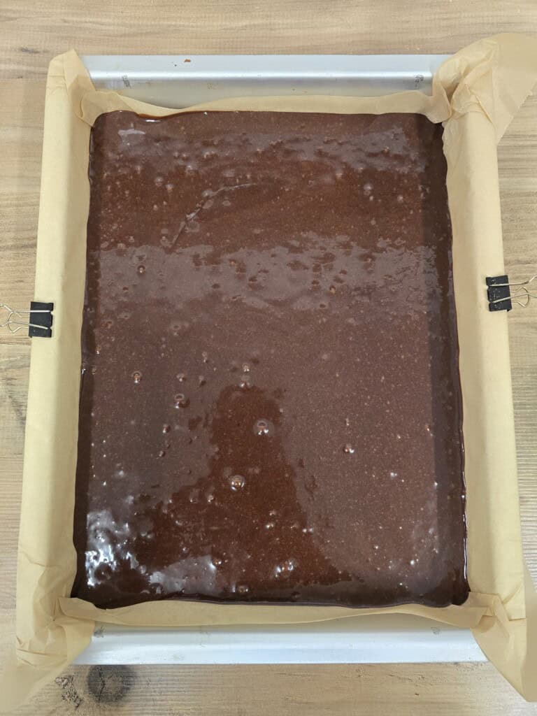 A rectangular baking tin lined with greaseproof paper is filled with unbaked chocolate creme egg brownie mixture, resting on a wooden surface.