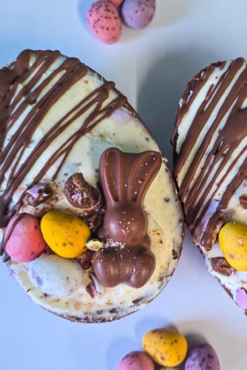 Two chocolate Easter egg halves filled with white chocolate, topped with a small chocolate bunny and colourful candy-coated eggs, sit on a white surface with more candy eggs scattered nearby.
