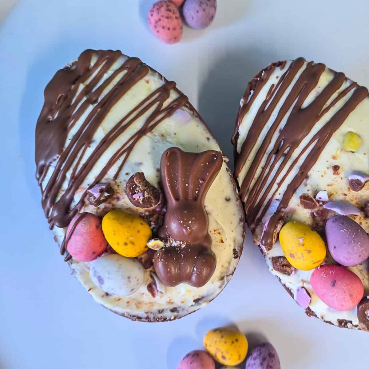 Two Easter Egg Cheesecake: Easter egg halves filled with cheesecake filling, topped with a small chocolate bunny and colourful candy-coated eggs, sit on a white surface with more candy eggs scattered nearby.