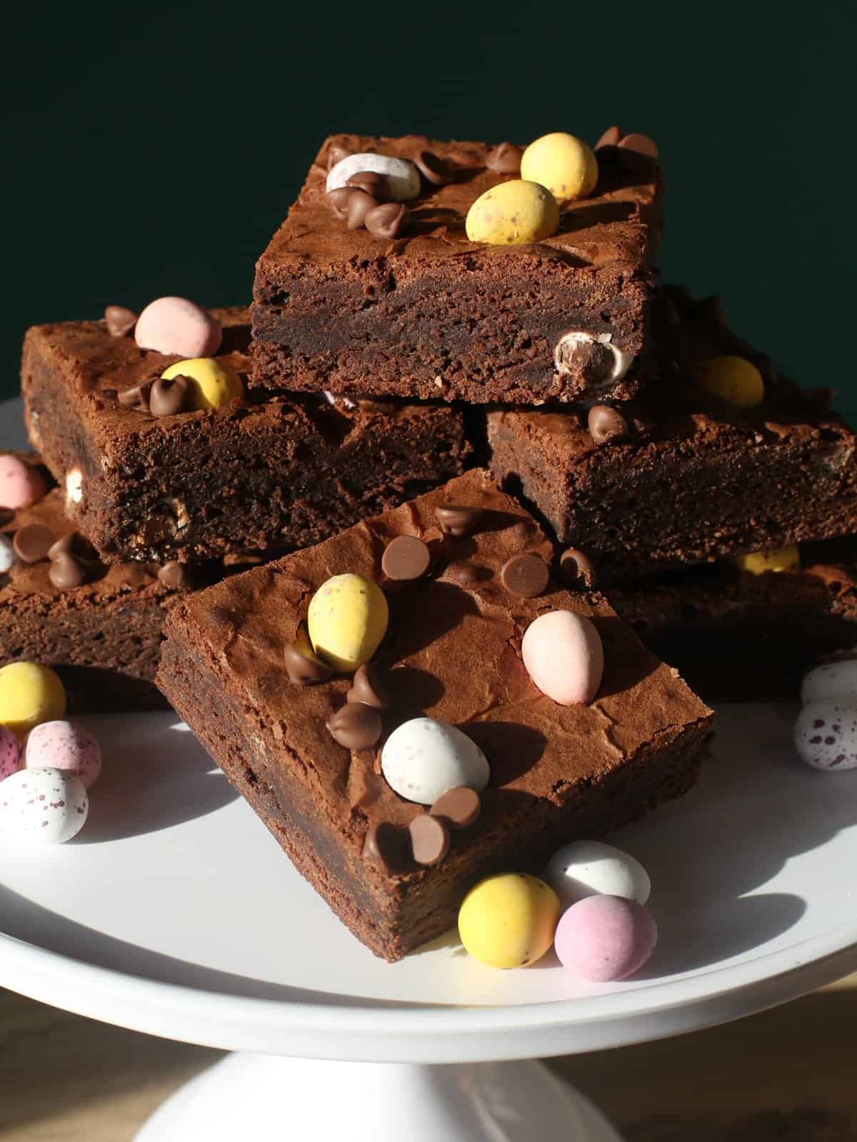 A stack of chocolate mini egg brownies topped with pastel-coloured candy eggs and chocolate chips, displayed on a white cake stand with more candy eggs scattered around.