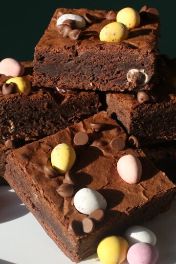 A stack of mini egg brownies topped with mini chocolate eggs and chocolate chips, arranged on a white plate with extra mini eggs scattered around.