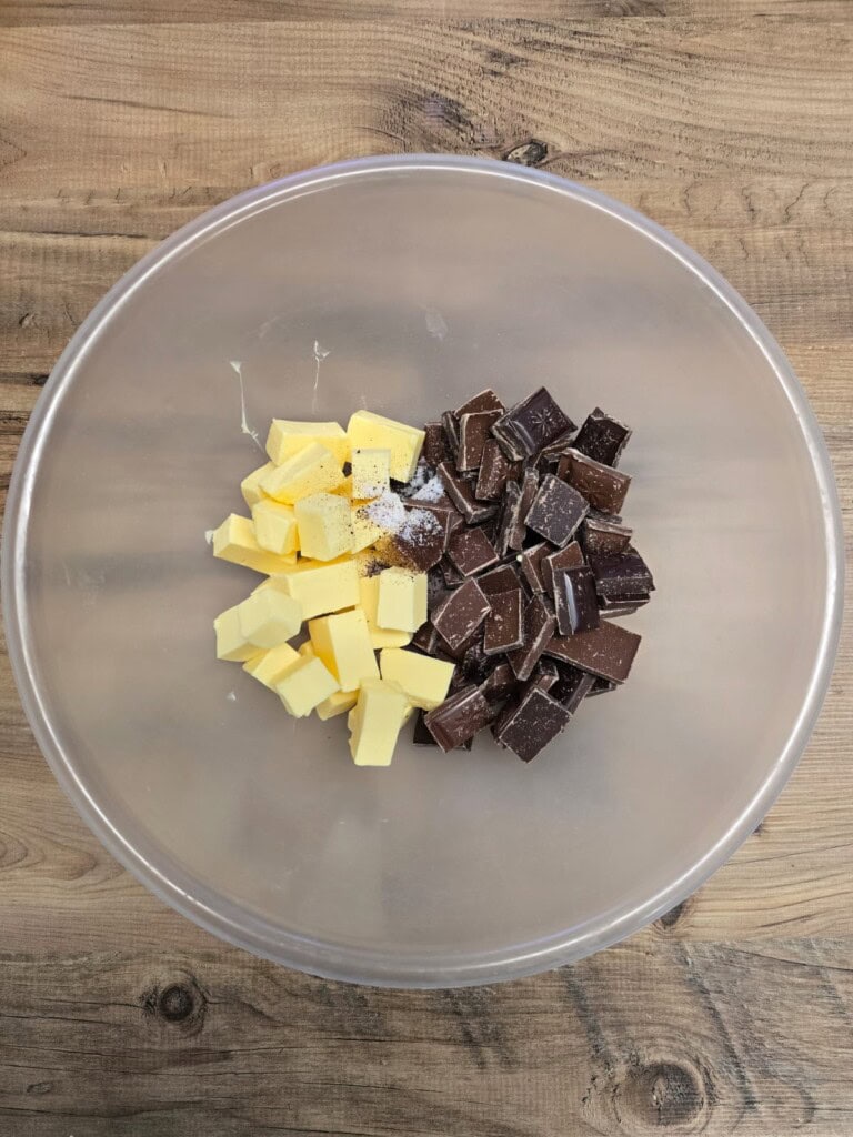 A clear mixing bowl on a wooden surface contains cubed butter and chopped dark chocolate, placed side by side.