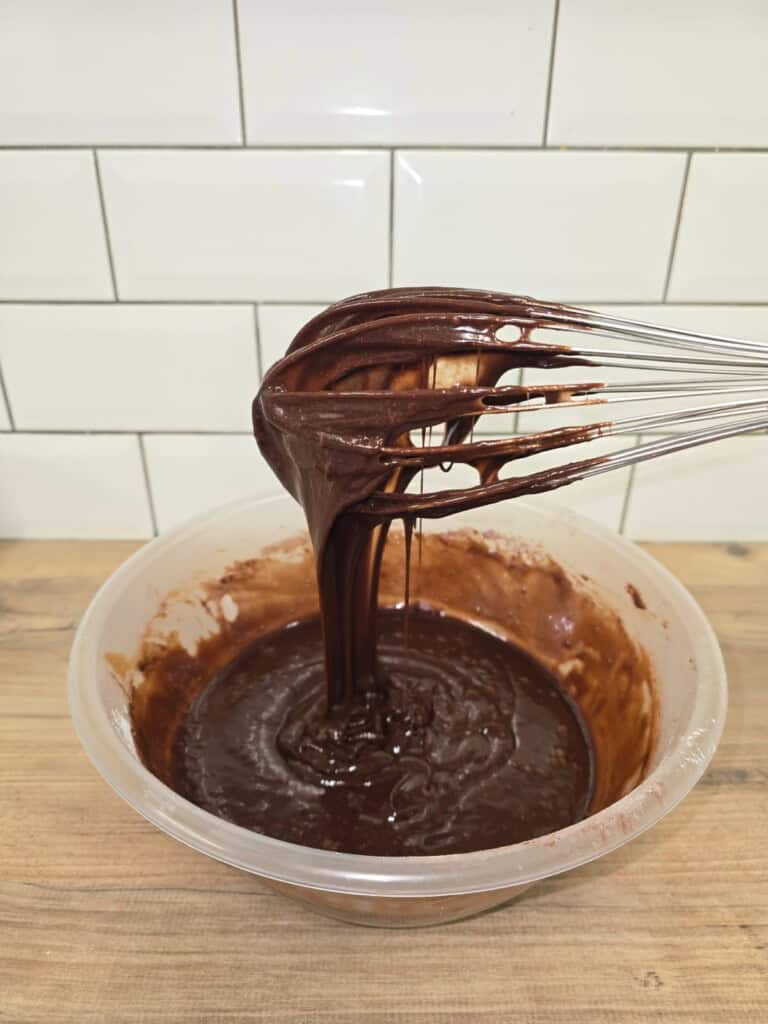 A metal whisk covered in chocolate cake mixture is held above a mixing bowl filled with more mixture, with a wooden worktop and white tiled wall in the background.