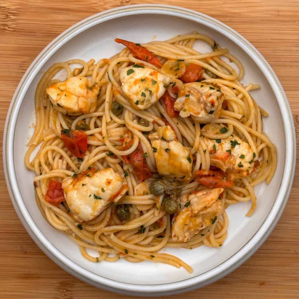 A plate of monkfish pasta: spaghetti topped with chunks of monkfish, diced tomatoes, capers, and chopped herbs, set on a wooden surface.