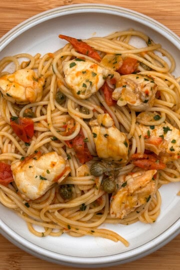 A plate of monkfish pasta: spaghetti topped with chunks of monkfish, diced tomatoes, capers, and chopped herbs, set on a wooden surface.