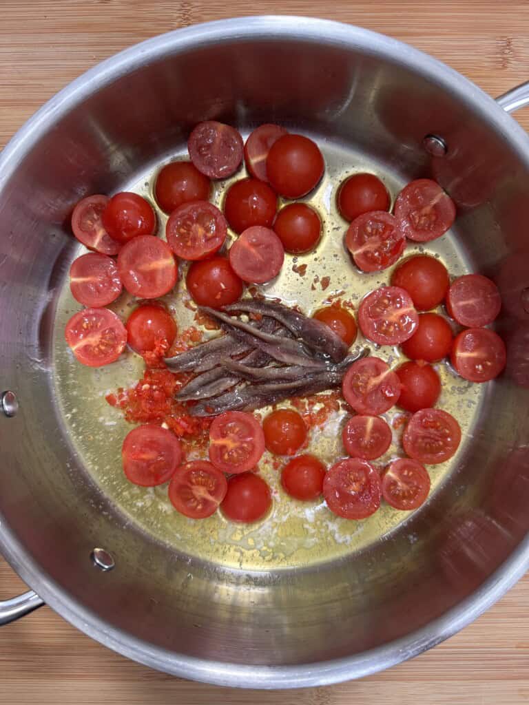A metal saucepan contains halved cherry tomatoes, several anchovy fillets, crushed chilli flakes, and olive oil on a wooden surface. The ingredients appear uncooked and are arranged in the saucepan.