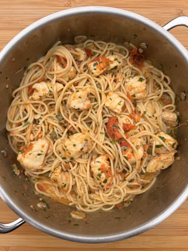 A large metal pot filled with monkfish pasta: cooked spaghetti, chunks of chicken, diced tomatoes, and herbs, sitting on a wooden surface.