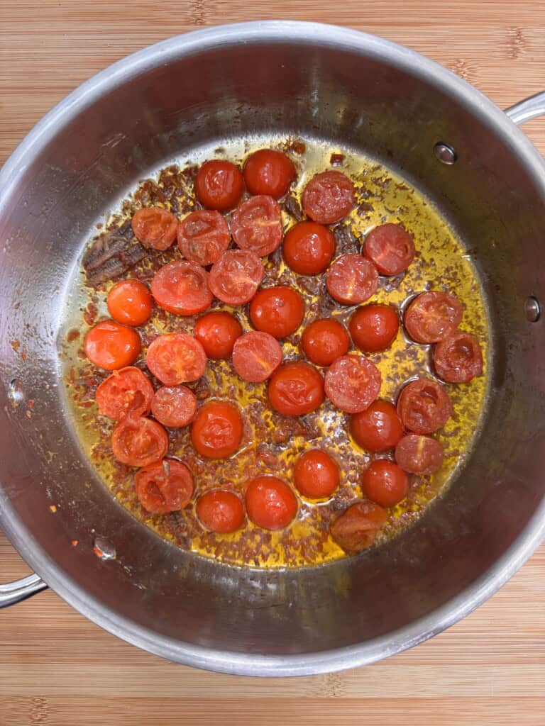 A stainless steel saucepan on a wooden surface contains sautéed cherry tomatoes, some halved and some whole, sizzling in oil with brown caramelised bits on the bottom.