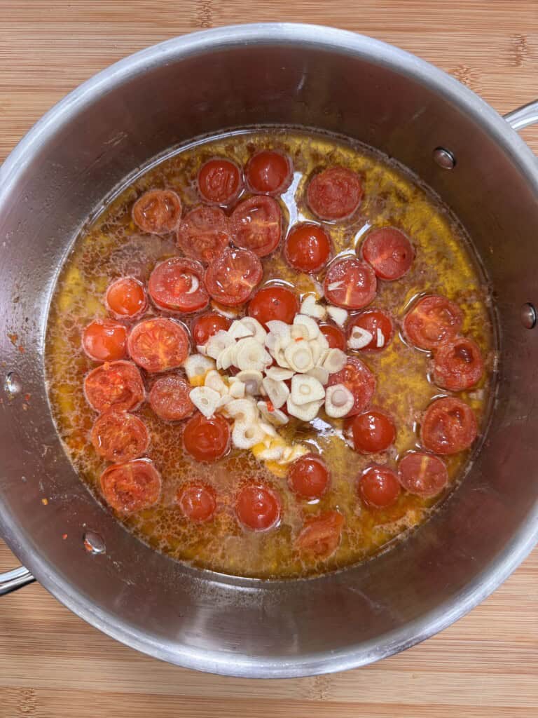 A stainless steel saucepan sits on a wooden surface, filled with halved cherry tomatoes, sliced garlic, and oil simmering together. The ingredients appear to be cooking, forming the base of a sauce.