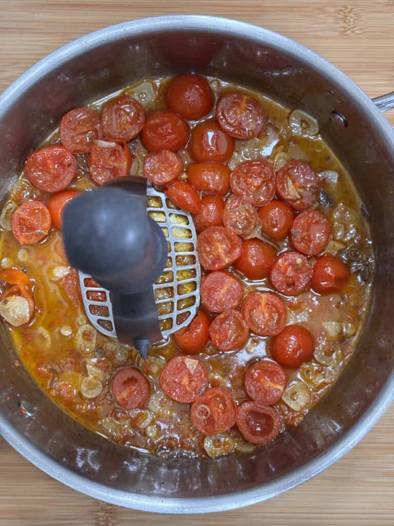 A top view of a saucepan with cherry tomatoes being mashed using a potato masher. The tomatoes are cooking in a sauce with visible sliced garlic and oil on a wooden surface.