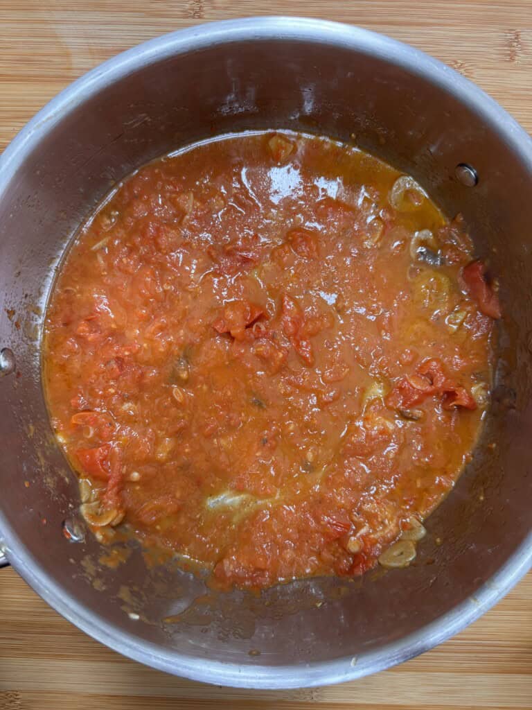 A stainless steel saucepan containing a chunky tomato sauce with visible tomato pieces and some oil, sitting on a wooden surface.