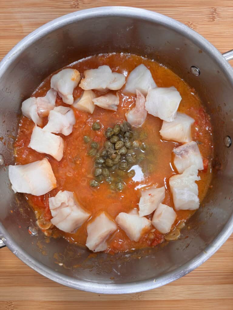 Chunks of raw white fish, capers, and a tomato-based sauce are in a stainless steel saucepan on a wooden surface, ready to be cooked.