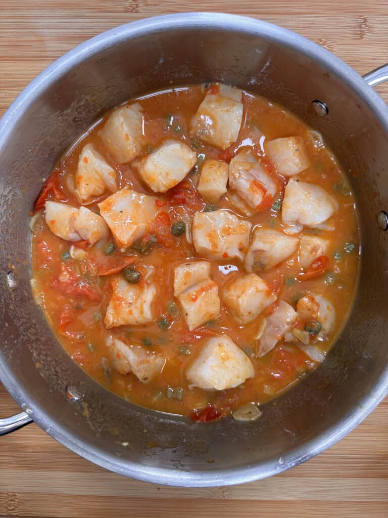 A stainless steel saucepan contains chunks of white fish in a tomato-based sauce with capers and diced vegetables on a wooden surface.