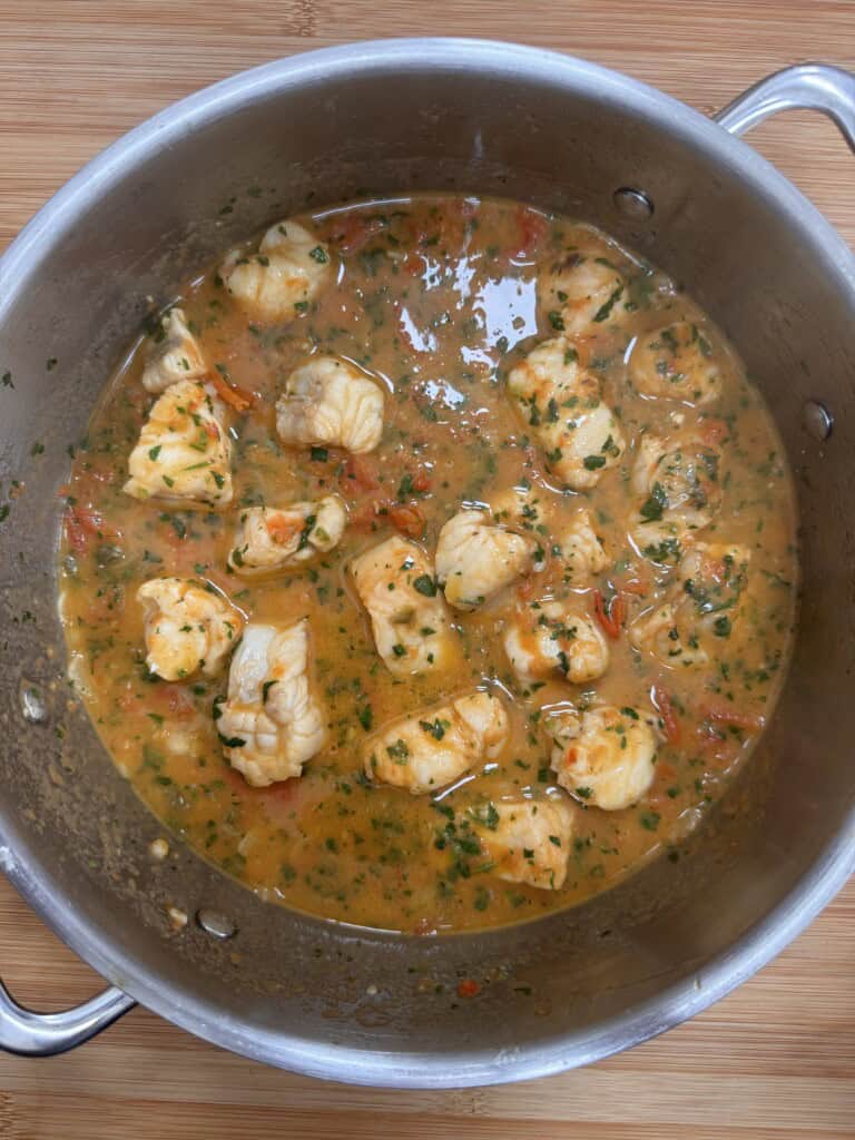 A pot filled with fish stew featuring chunks of white fish in an orange, tomato-based broth with fresh herbs and pieces of tomato, set on a wooden surface.