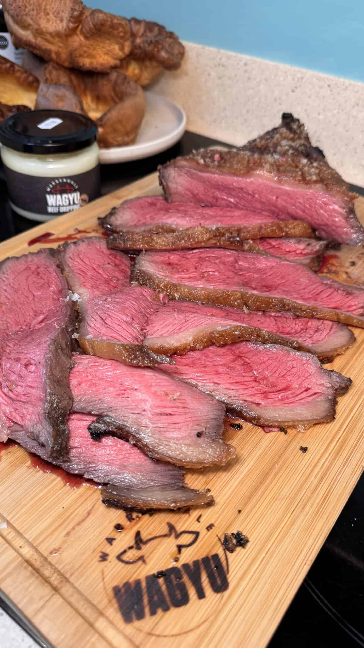 Sliced medium-rare Wagyu beef is displayed on a wooden chopping board, showing a juicy pink centre and browned edges. A jar labelled “WAGYU” and a plate with food are in the background on a kitchen worktop.
