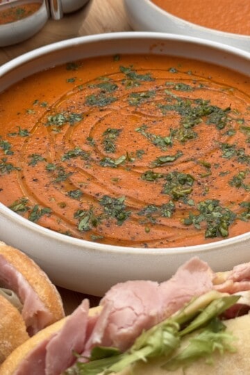 A bowl of creamy zaatar roasted red pepper and tomato soup garnished with herbs and black pepper, with ham sandwiches on crusty bread in the foreground and another bowl of soup in the background.