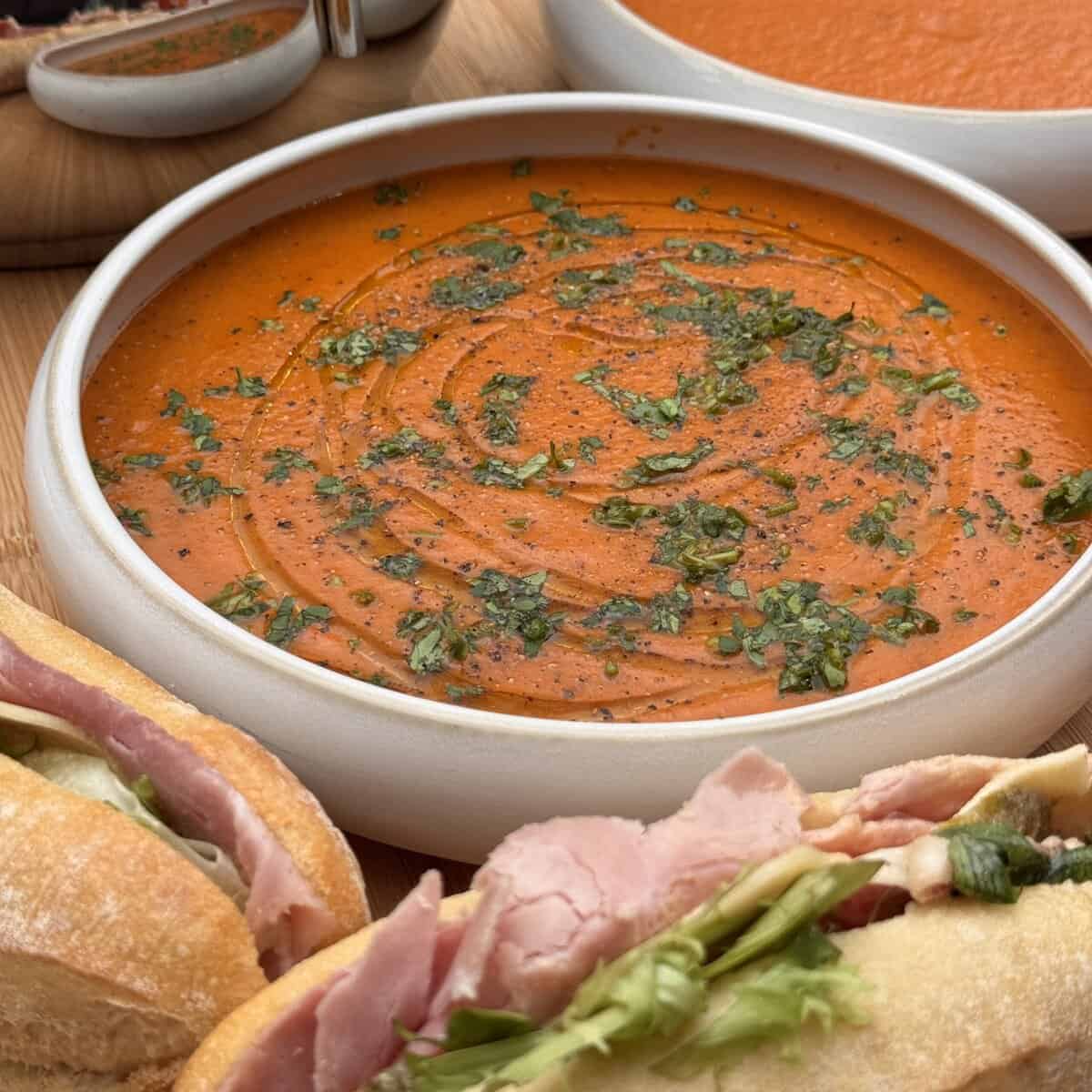 A bowl of creamy zaatar roasted red pepper and tomato soup garnished with herbs and black pepper, with ham sandwiches on crusty bread in the foreground and another bowl of soup in the background.