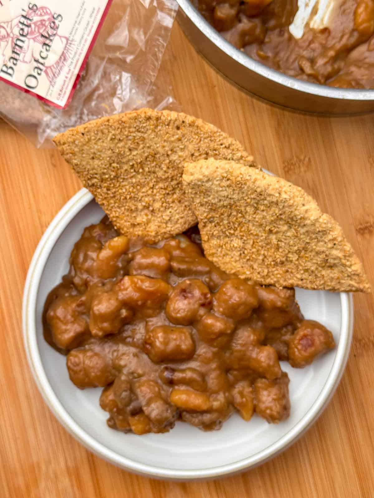 A bowl of hearty Scottish stovies with chunks of meat and two large, triangular oatcakes on top, placed on a wooden surface. A packet of oatcakes and a pan of stew are partially visible in the background.