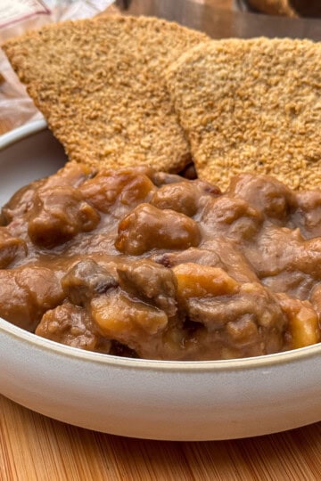 A bowl of thick Scottish stovieswith visible chunks of meat and vegetables, served with two large, crispy wholegrain crackers on a wooden surface.